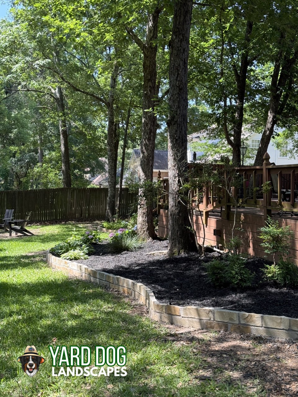Long curved East Texas flower bed with fresh black mulch and sandstone block edging running between a deck and privacy fence