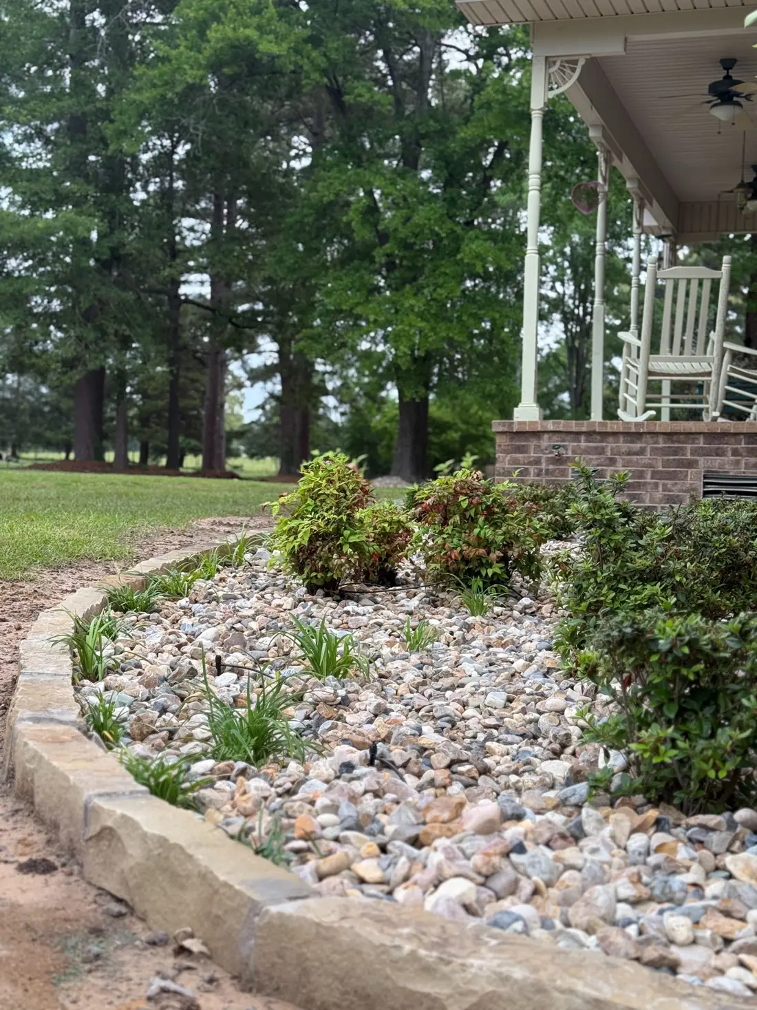 Low-water planting bed with river rock, monkey grass, and evergreen shrubs bordering a brick home with a white porch, built to hold up through the East Texas summer heat