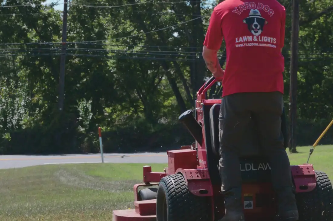 A Yard Dog crew member in a red shirt running a red stand-on mower along a green roadside strip, grass clippings spraying out the side